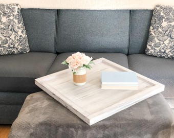 May include: A white wooden tray with a raised edge sits on a gray ottoman. The tray has two books and a small vase with pink flowers on it.