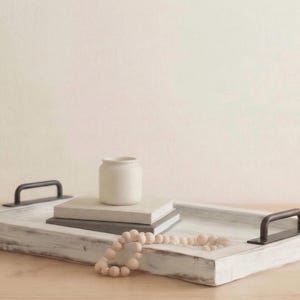 May include: A white distressed wooden tray with black metal handles, holding a small white vase, books, and a beaded garland. The tray is on a light wood surface.