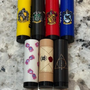 May include: Seven cylindrical objects with black bases. The tops are yellow, black, red, and blue, each with a different crest. The bottom row includes a white cylinder with pink designs, a tan cylinder with a scroll design, and a black cylinder with a triangle design.