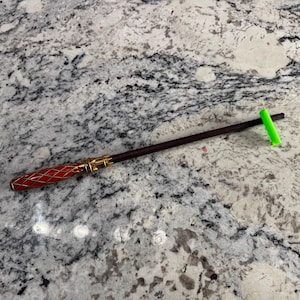 May include: A dark brown wooden wand with a red and gold handle. The wand is lying on a white countertop with a green marker next to it.