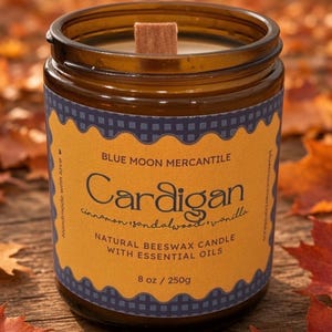 Peut inclure: Une bougie en cire d'abeille naturelle dans un pot en verre brun avec une mèche en bois. L'étiquette présente un motif festonné jaune sur fond à carreaux bleu, indiquant Blue Moon Mercantile, Cardigan, cinnamon, sandalwood, vanilla, 250g.