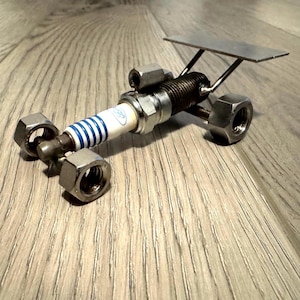 以下が含まれることがあります： 手作りの金属製飛行機彫刻。本体は青いストライプとフォードのロゴが入ったスパークプラグです。翼は金属製で、ナットとボルトで固定されています。彫刻は木製の表面にあります。