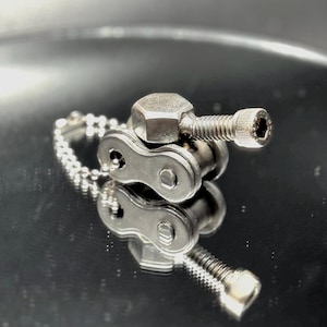 May include: A silver metal keychain shaped like a tank, made from a bike chain and a bolt. The keychain has a silver chain attached to it.
