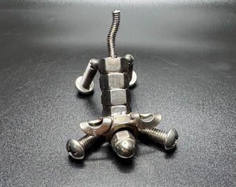 May include: A metal sculpture of a person constructed from nuts, bolts, and a spring. The figure features a body of stacked nuts, arms of bolts, and a spring for a neck. The head is a nut, and the feet are bolts.