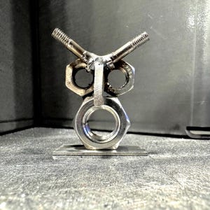Può includere: Una scultura di gufo in metallo realizzata con dadi, bulloni e una piastra di metallo. Gli occhi del gufo sono formati da dadi esagonali, con bulloni come sopracciglia. Il corpo è fatto di un dado grande e la scultura è montata su una base metallica rettangolare.