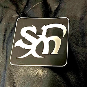May include: A black square sticker with a white stylized "SH" design. The letters are intertwined with decorative flourishes. The sticker has a white border and is set against a dark, textured background.