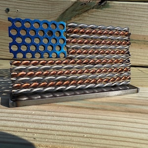 May include: A metal sculpture of the American flag. The blue section features a perforated design, while the stripes are made of twisted metal in silver and copper colors. The sculpture sits on a rectangular base.