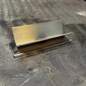 May include: A rectangular metal stand featuring a flat base and an angled upright plate. The object is constructed from steel with visible heat discoloration, showing shades of gold, bronze, and silver across the metallic surfaces.