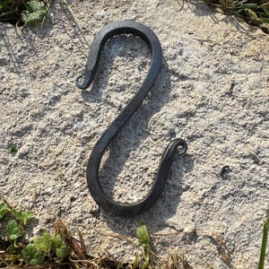 Könnte beinhalten: Handgeschmiedeter S-Haken aus dunklem Metall. Der Haken ist gebogen und hat an jedem Ende eine spiralförmige Verzierung. Der Haken wird auf einer rauen, grauen Steinoberfläche mit grünem Gras und kleinen Pflanzen im Hintergrund gezeigt. Ein dekorativer und funktioneller Artikel.