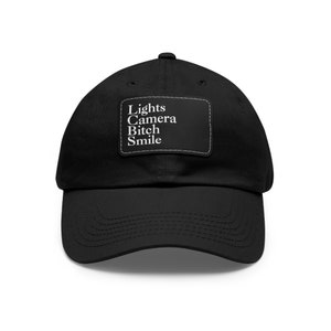May include: Black baseball cap with a curved peak. A rectangular patch on the front displays the words "Lights Camera Bitch Smile" in white, stitched onto the cap. The cap is made of a soft fabric.