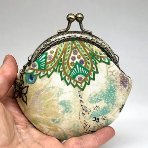 Könnte beinhalten: Eine kleine, runde Münzbörse mit einem Messingverschluss und einem Stoff mit Blumenmuster. Der Stoff zeigt ein Paisley-Muster in Grün-, Gold- und Weißtönen. Die Börse hat einen kleinen Schmetterlingsanhänger am Verschluss.