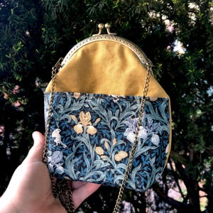Könnte beinhalten: Kleine Umhängetasche mit senfgelbem Oberteil und floral bedruckter Basis. Die Tasche hat einen Metallverschluss und einen Kettenriemen. Das Blumenmuster ist dunkelblau mit weißen und goldenen Blumen und Blättern.