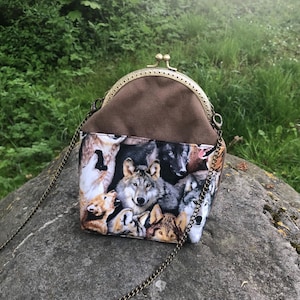 Könnte beinhalten: Kleine Umhängetasche mit Wolfsdruck. Die Tasche hat einen Metallverschluss und einen Kettenriemen. Der Wolfsdruck zeigt verschiedene Wolfsgesichter in Grau-, Braun- und Weißtönen. Der obere Teil der Tasche ist aus braunem Stoff.