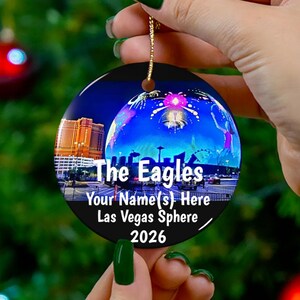 May include: A black round Christmas ornament with the text "The Eagles" and "Your Name(s) Here Las Vegas Sphere 2026". The ornament features a nighttime cityscape with fireworks and a sphere.