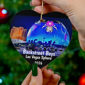 May include: Heart-shaped Christmas ornament featuring the Backstreet Boys Las Vegas Sphere 2026. The ornament displays a vibrant image of the Las Vegas Sphere at night. The ornament is held against a backdrop of a Christmas tree with red ornaments.