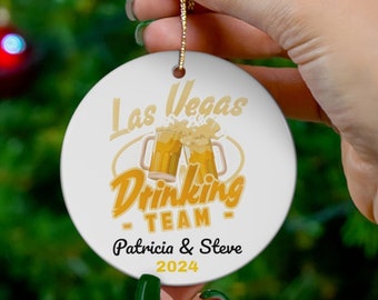 May include: White ceramic Christmas ornament with a gold design. The ornament features the text "Las Vegas Drinking Team" with two beer mugs and the names "Patricia & Steve" and the year "2024".