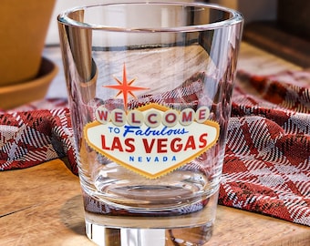 May include: A clear glass with a red, yellow, and blue graphic that says "Welcome to Fabulous Las Vegas, Nevada". The glass is sitting on a wooden cutting board with a red and white checkered cloth.