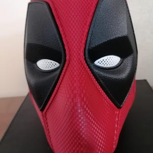 May include: A full-head mask featuring a textured red surface with a distinct grid pattern. The mask includes large, black, smooth eye patches surrounding white, mesh-like eye lenses. The design covers the entire face and head area.