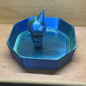 May include: A 3D-printed, iridescent blue and green decorative tray with a stylized bird figurine in the center. The tray has an octagonal shape with a textured surface, and the bird has a pointed beak.