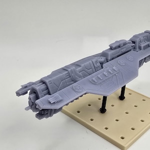 May include: A gray, futuristic spaceship model with intricate details, mounted on a beige base with black supports. The model features a long, streamlined body with various protrusions and a detailed front section, suggesting a science fiction theme.