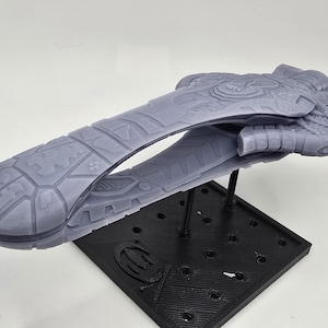 May include: A gray, detailed model of a spaceship, mounted on a black stand. The spaceship has intricate surface details and a sleek, elongated design. The stand is square with a logo.