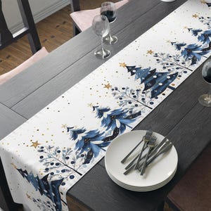 May include: A white table runner with a blue Christmas tree design, gold stars, and berries. The runner is placed on a dark wood table set for a holiday meal. Wine glasses, plates, and silverware are also on the table.