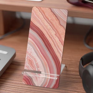 May include: A rectangular phone stand with a pink and red agate stone pattern. The stand is made of wood and has a clear plastic support. The stand is on a wooden desk with a keyboard, headphones, and other items.