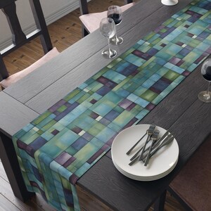May include: A rectangular table runner with a geometric pattern in shades of teal, blue, green, and purple. The runner is placed on a dark wooden table set for a meal, with plates, silverware, and wine glasses.