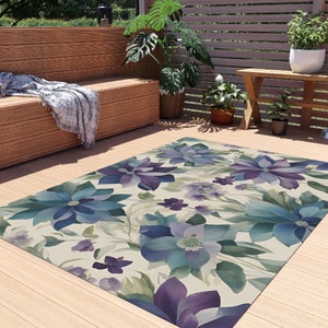 May include: A floral patterned outdoor rug with blue and purple flowers on a cream background. The rug is laid out on a wooden deck.