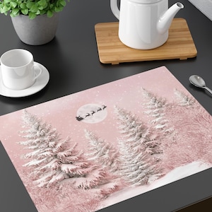 May include: A rectangular placemat with a winter scene featuring snow-covered trees and a full moon with Santa's sleigh. The placemat has a pink background and white accents, creating a festive holiday theme.