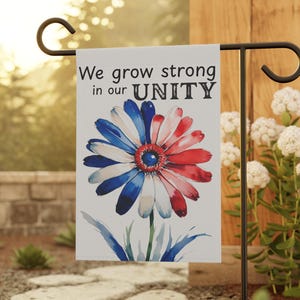 May include: A garden flag featuring a watercolour flower design in red, white, and blue, with the words "We grow strong in our UNITY." The flag is displayed on a black metal stand in a garden.