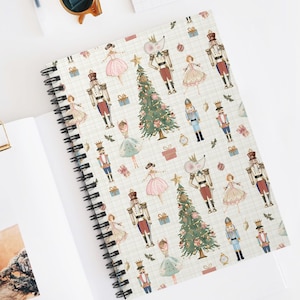Peut inclure: Un carnet à spirales avec un motif festif sur le thème de Casse-Noisette. La couverture présente des illustrations de soldats Casse-Noisette, de ballerines, de sapins de Noël et de boîtes cadeaux sur un fond quadrillé. Le carnet est ouvert sur une page blanche.