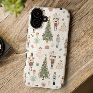 Może przedstawiać: Etui na telefon z świątecznym wzorem bożonarodzeniowym. Projekt przedstawia ilustracje dziadków do orzechów, choinek, myszy i baletnic w powtarzającym się wzorze na białym tle. Etui leży na drewnianej powierzchni.