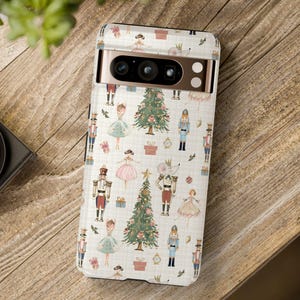 Puede incluir: Funda de teléfono con un tema festivo de Cascanueces. La funda presenta un patrón repetido de bailarinas, soldados de juguete, árboles de Navidad y regalos sobre un fondo crema. La funda está sobre una superficie de madera.