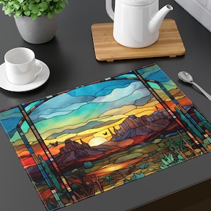 May include: A rectangular placemat featuring a stained-glass style desert landscape with a sunset. The design includes mountains, cacti, and a vibrant sky with shades of blue, orange, and yellow. The placemat is on a dark surface.