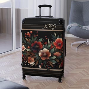 May include: A black rolling suitcase featuring a floral pattern with red, orange, and cream-colored flowers and green leaves. The luggage has a retractable handle and the letters KRS printed in a cursive white font near the top edge.
