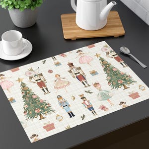 May include: A rectangular placemat with a festive Nutcracker theme. The cream-colored mat features illustrations of nutcrackers, ballerinas, Christmas trees, and wrapped gifts. The design includes a grid pattern and is suitable for holiday dining.