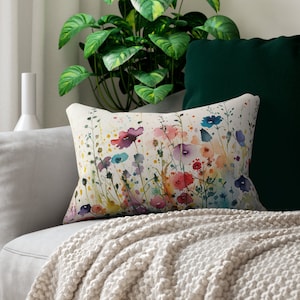 Könnte beinhalten: Ein rechteckiges Zierkissen mit einem Aquarelldruck von Wildblumen in Rosa-, Blau-, Lila- und Gelbtönen auf cremefarbenem Hintergrund. Das Design zeigt zarte Stängel, Blätter und Farbspritzer auf der Oberfläche des Stoffes.