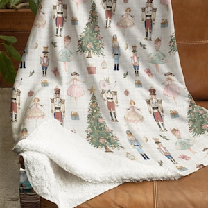 May include: A cream-colored throw blanket with a repeating pattern of nutcrackers, ballerinas, Christmas trees, and gifts. The blanket has a soft, white sherpa lining. The blanket is draped over a brown leather couch.