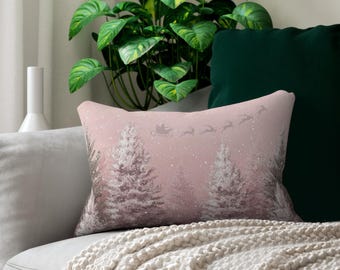 Könnte beinhalten: Ein rechteckiges Dekokissen mit einem zartrosa Hintergrund und einer winterlichen Waldszene. Das Design zeigt weiße, schneebedeckte Tannenbäume und die Silhouette eines von Rentieren gezogenen Schlittens am Himmel, akzentuiert durch weiße Schneeflocken.