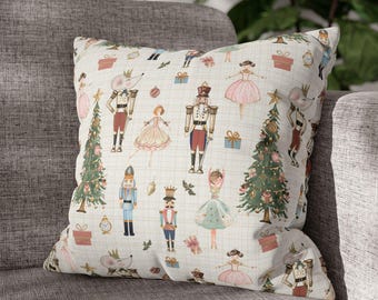 May include: Square decorative pillow with a whimsical Nutcracker theme. The off-white pillow features a repeating pattern of Nutcracker soldiers, ballerinas, Christmas trees, mice, and gift boxes. The pillow is resting on a gray sofa.