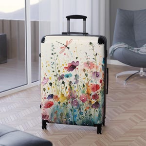 May include: A hard-shell rolling suitcase featuring a cream-colored background with a vibrant watercolor print of various wildflowers, green stems, and a single dragonfly. The luggage includes a black retractable handle and four black spinner wheels.