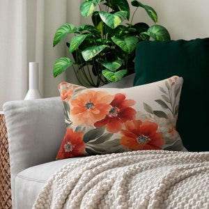 May include: A rectangular throw pillow featuring a watercolor-style floral print with large orange and red blossoms set against a light cream background, accented by muted green foliage and stems, displayed on a light gray upholstered sofa.