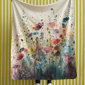 May include: A white throw blanket with a watercolour floral print featuring pink, purple, blue, and yellow flowers. The blanket is being held up by a person, showing the full design.