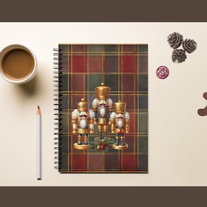 May include: A spiral-bound notebook featuring a dark red and green plaid pattern with a central illustration of three gold nutcracker figures. The notebook is accompanied by a cup of coffee, a pencil, pinecones, a candy cane, and a gingerbread man.