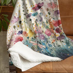 May include: A white throw blanket with a watercolor design of colorful flowers and a dragonfly. The blanket has a soft, fuzzy texture.