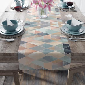 May include: A geometric patterned table runner in shades of blue, peach, and gray. The runner is placed on a wooden table set for a meal, with plates, bowls, silverware, and wine glasses. A vase of pink flowers sits in the center.