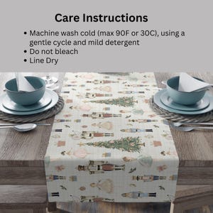 May include: A cream-colored table runner with a repeating pattern of nutcrackers, Christmas trees, and gift boxes. The runner is displayed on a wooden table, with place settings on either side. Care instructions are printed at the top.