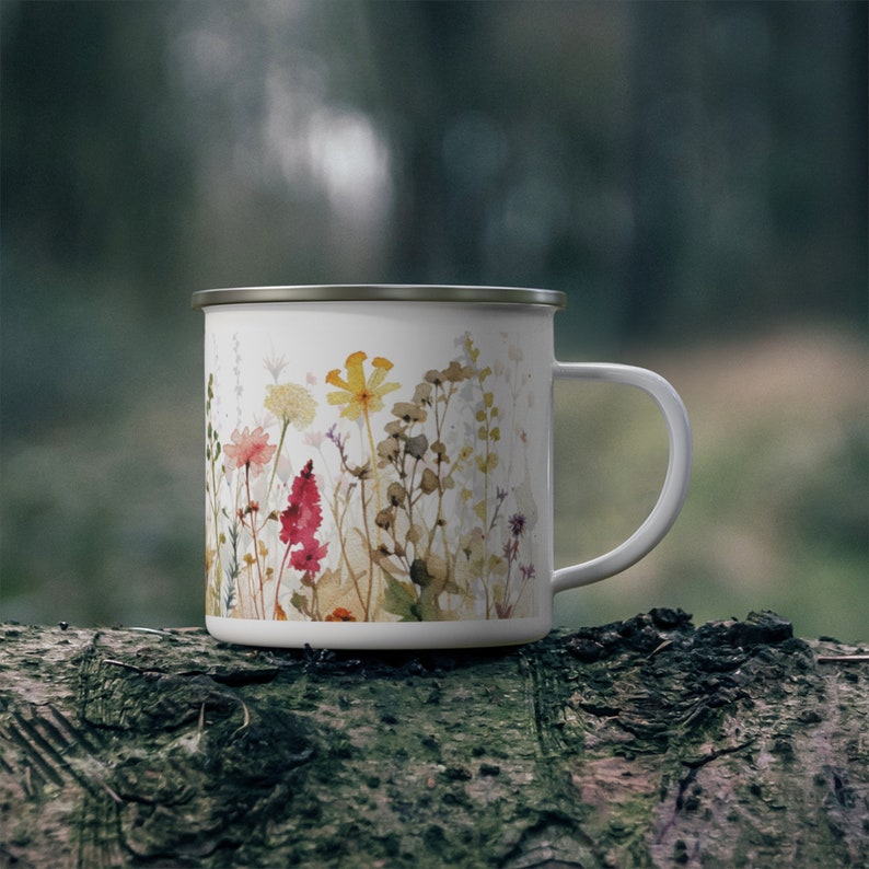May include: A white enamel mug featuring a colorful watercolor-style print of various wildflowers in shades of yellow, pink, and brown. The mug has a smooth white finish, a simple handle on the right side, and a metallic silver rim along the top edge.