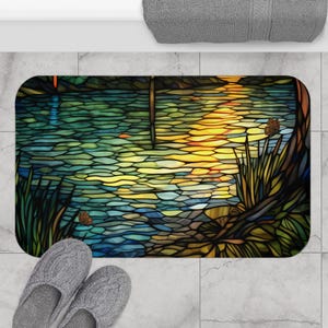 Puede incluir: Una alfombra de ba&ntilde;o de vidrio tintado con un dise&ntilde;o de un lago con una puesta de sol reflejada en el agua. La alfombra tiene un borde marr&oacute;n oscuro y una variedad de colores, incluyendo azul, verde, amarillo y naranja.